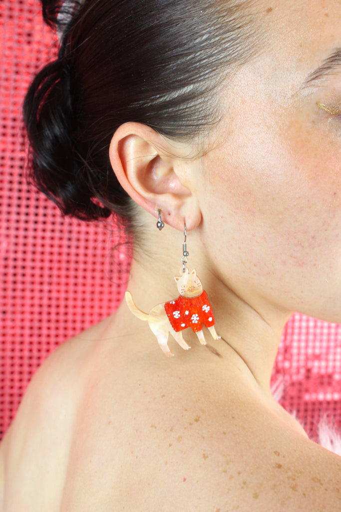 Model wearing earrings that feature acrylic ginger cats with red Christmas jumpers that have snowflakes on them, hanging from stainless steel earwires