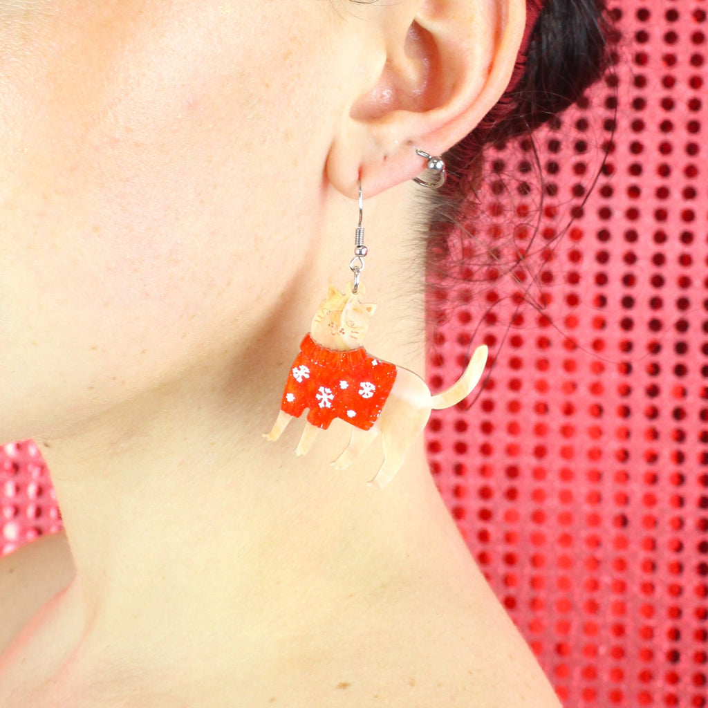 Model wearing earrings that feature acrylic ginger cats with red Christmas jumpers that have snowflakes on them, hanging from stainless steel earwires