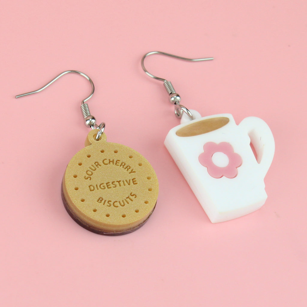 Chocolate digestive charm reading 'Sour Cherry Digestive Biscuits' and a cup of tea charm with a pink flower design, both on stainless steel earwires