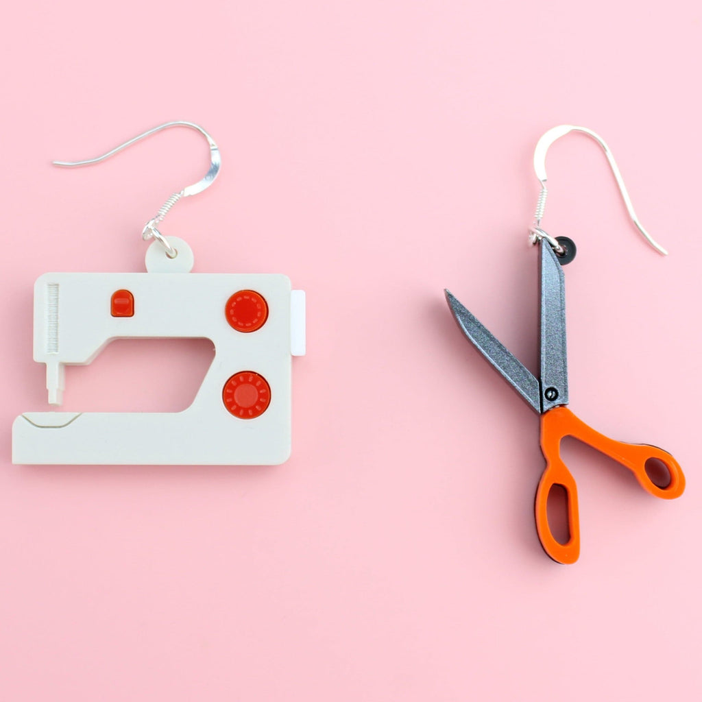 White and red sewing machine charm and a scissor charm with an orange handle, both on stainless steel earwires
