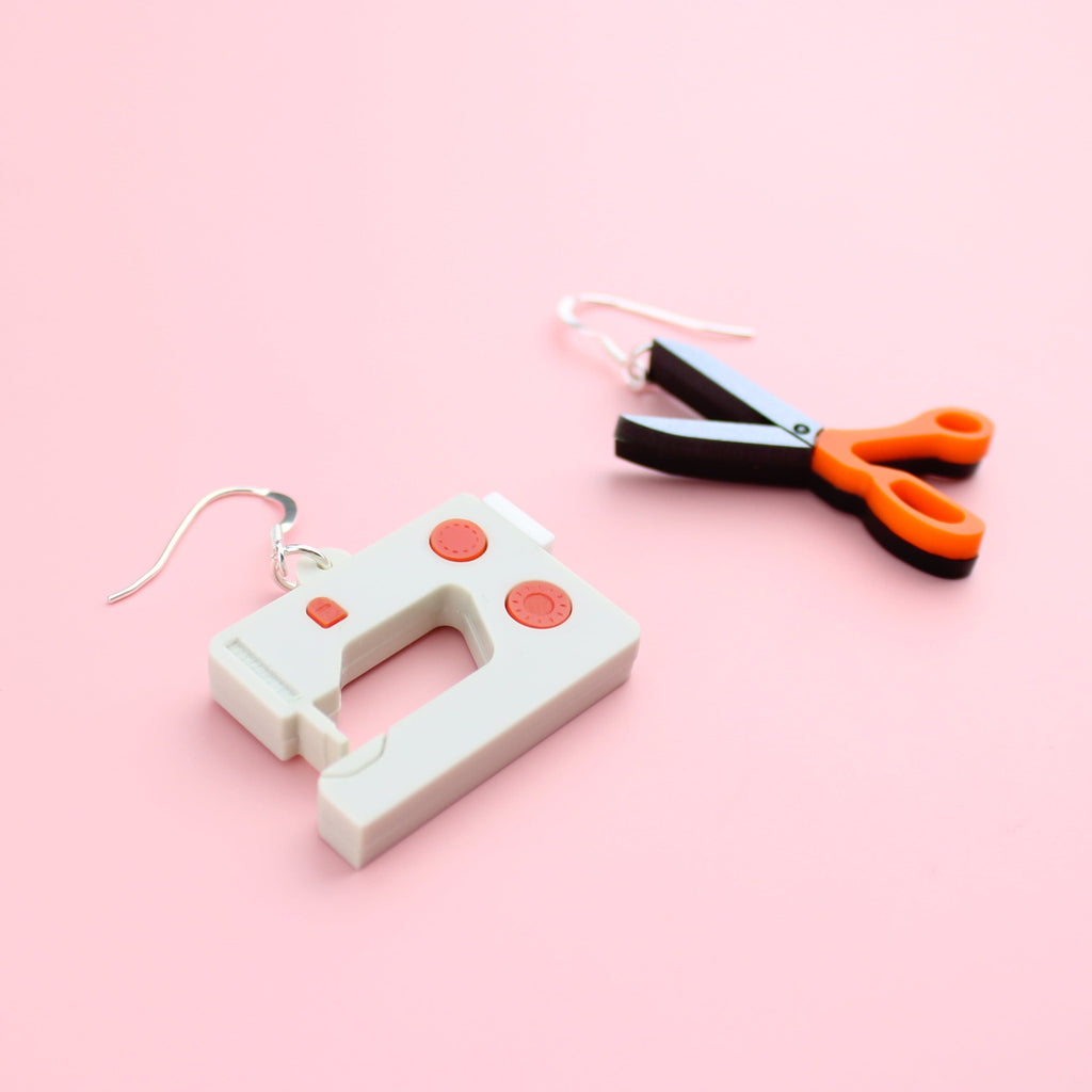 White and red sewing machine charm and a scissor charm with an orange handle, both on stainless steel earwires